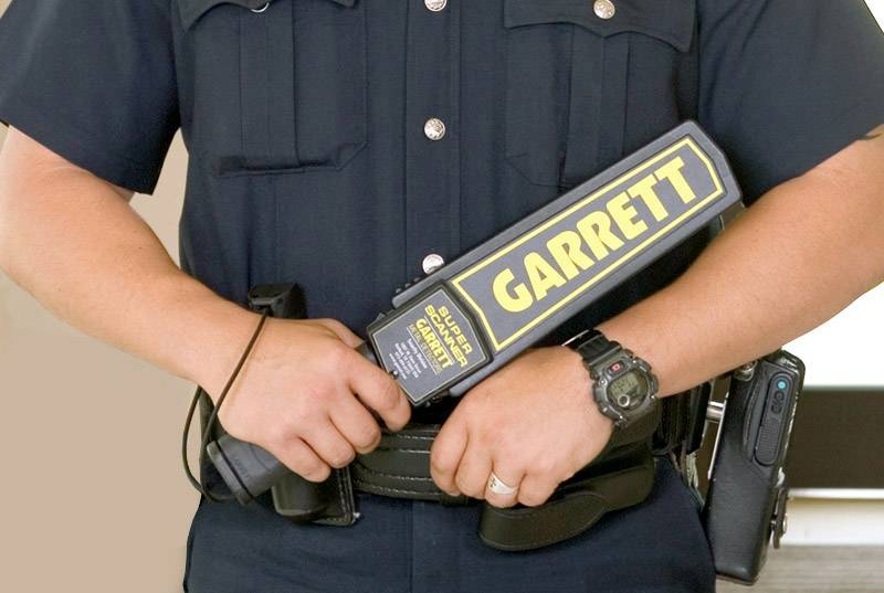 Bezpečnostný pracovník drží ručný kovový detektor s nápisom 'GARRETT'. Má na sebe čiernu uniformu s príslušenstvom na opasku.