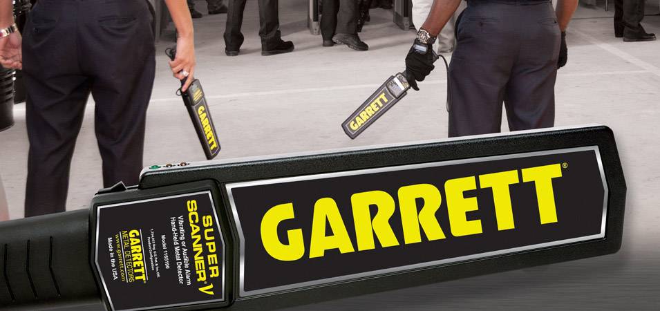 Niekoľko bezpečnostných pracovníkov používa ručné detektory kovov na kontrolu osôb. Nápis 'Garrett' je dobre viditeľný na zariadeniach.