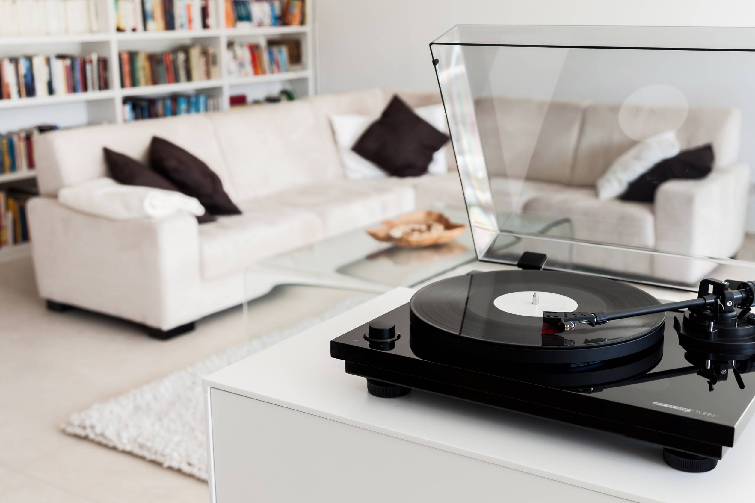 Gramofón s čiernou vinylovou platňou v popredí modernej obývacej izby s béžovými sedačkami a knižnicou v pozadí.