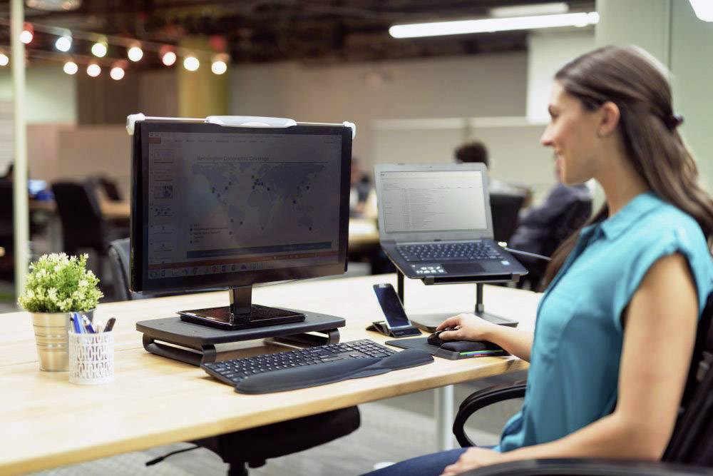 Žena pracuje pri stole s dvoma monitormi. Na väčšom monitore je viditeľná mapa sveta.
