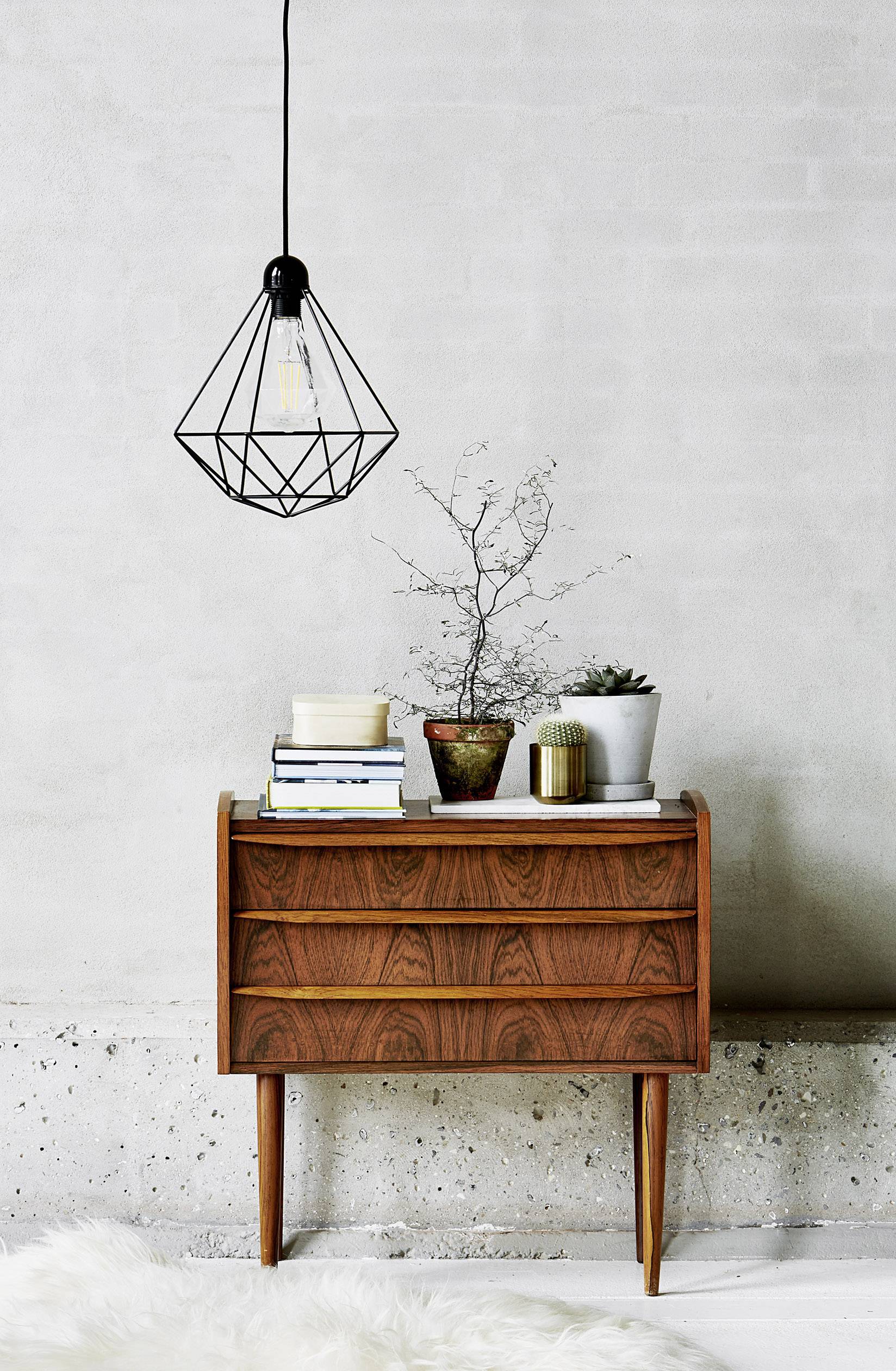 Malá drevená skriňa s tromi zásuvkami stojí pred sivou stenou. Na skrini sú umiestnené rastliny, knihy a sviečky. Moderné závesné svetlo visí nad ňou.