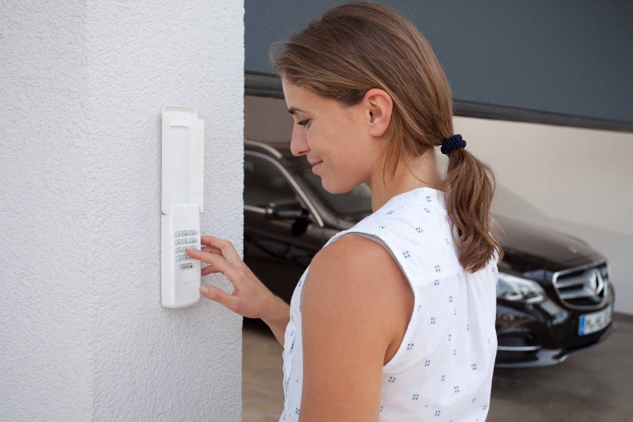 Žena stláča tlačidlá na zariadení na stene, pravdepodobne vrátový telefón alebo bezpečnostný kód; v pozadí je automobil.