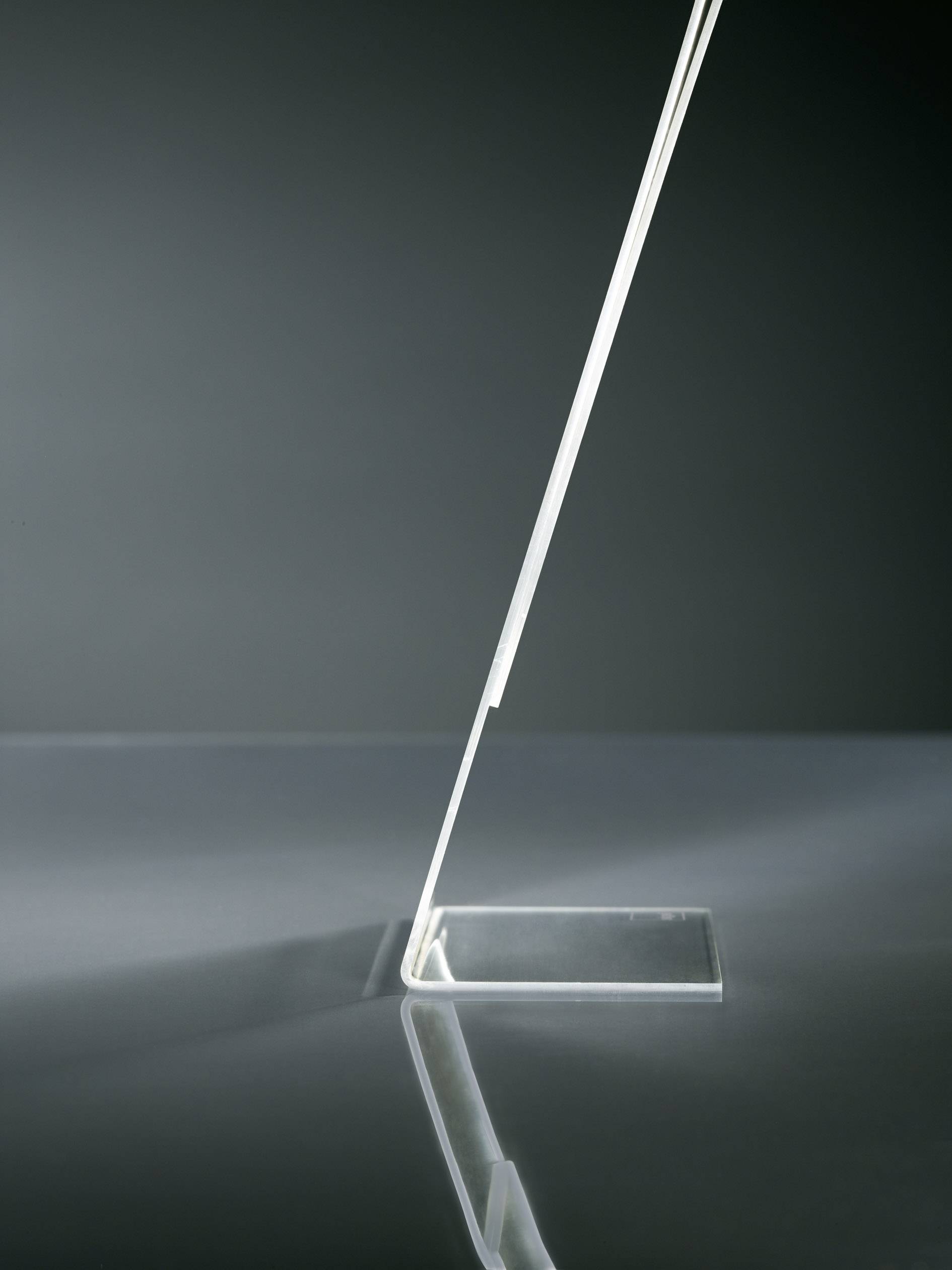 Úzka, moderná noha stolnej lampy z priehľadného materiálu, elegantne naklonená, odráža sa na lesklom povrchu.
