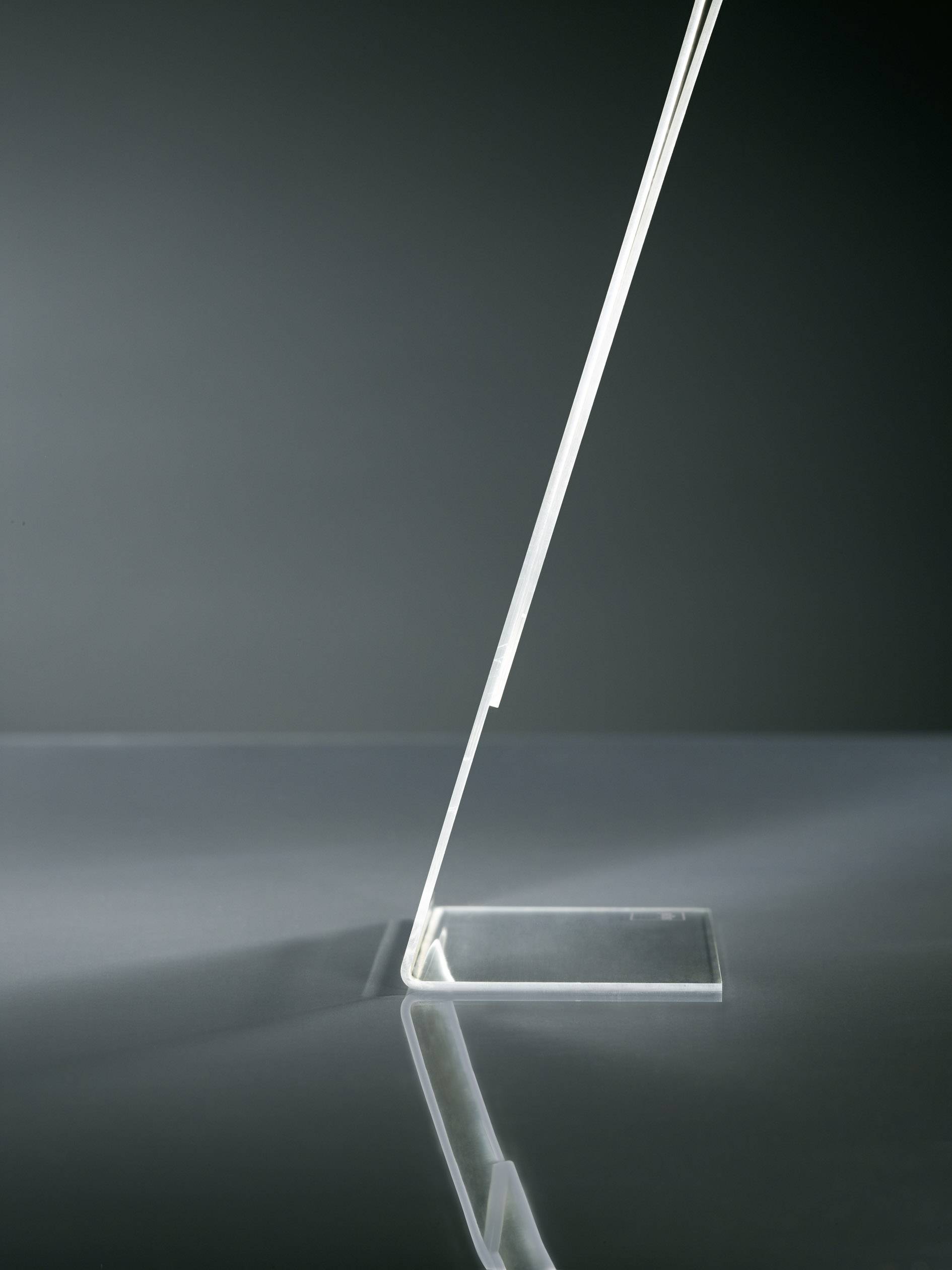 Minimalistická, moderná stolná lampa s tenkým, obdĺžnikovým podstavcom a dlhým, šikmo smerujúcim stĺpikom na sivom povrchu.