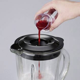 Jedna ruka nalieava červenú tekutinu z malého pohára do skleného mixéra s čiernym vekom, ktorý má dole biely obsah.