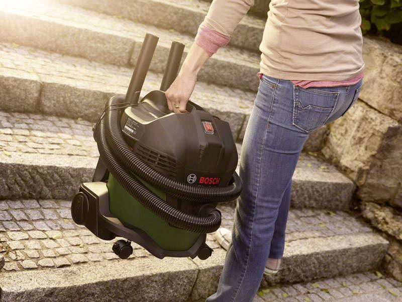 Osoba nesie priemyselný vysávač Bosch do schodiska, vysávač má flexibilnú hadicu a kolieskа.