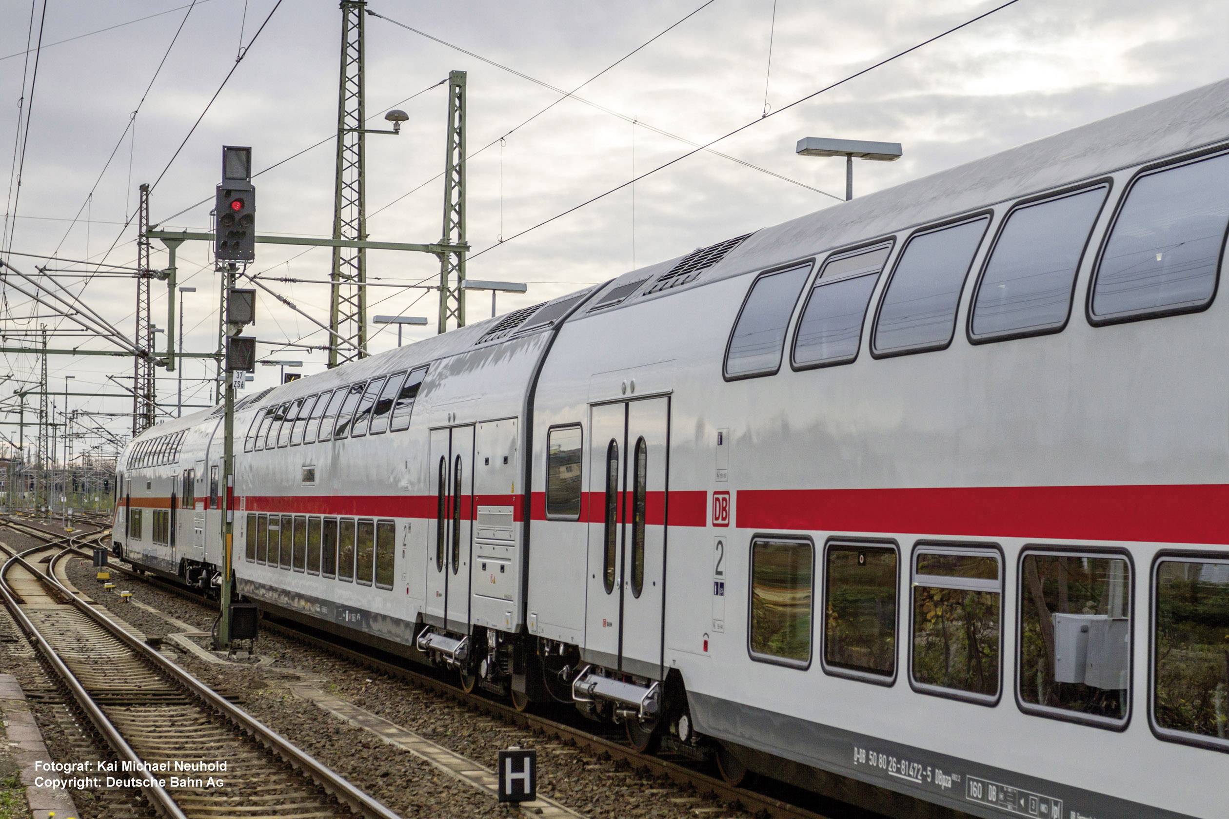 Strieborný dvojposchodový vlak Nemeckých dráh (Deutsche Bahn) jazdí po koľaji. Vedľa neho stoja signálne stožiare a trakčné vedenia na železničnej stanici.
