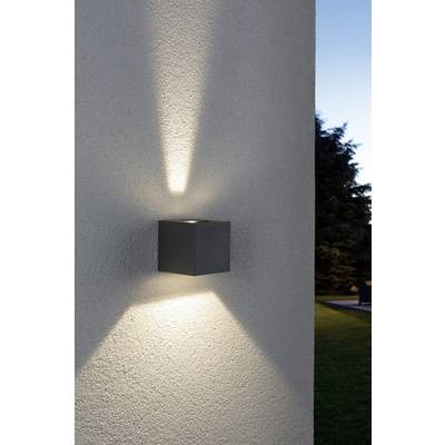 Nástenná lampa na bielej vonkajšej stene so žiariacim svetelným kužeľom smerujúcim nahor a nadol. V pozadí stromy a modrá obloha večer.