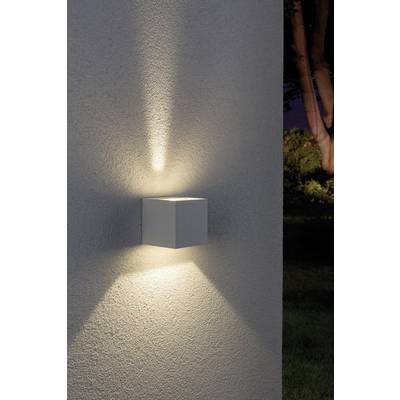 Vonkajšia nástenná lampa v podobe bieleho kockovitého tvaru, ktorá vyžaruje svetlo smerom nahor a nadol na betónovej stene, obklopená stromami v pozadí.