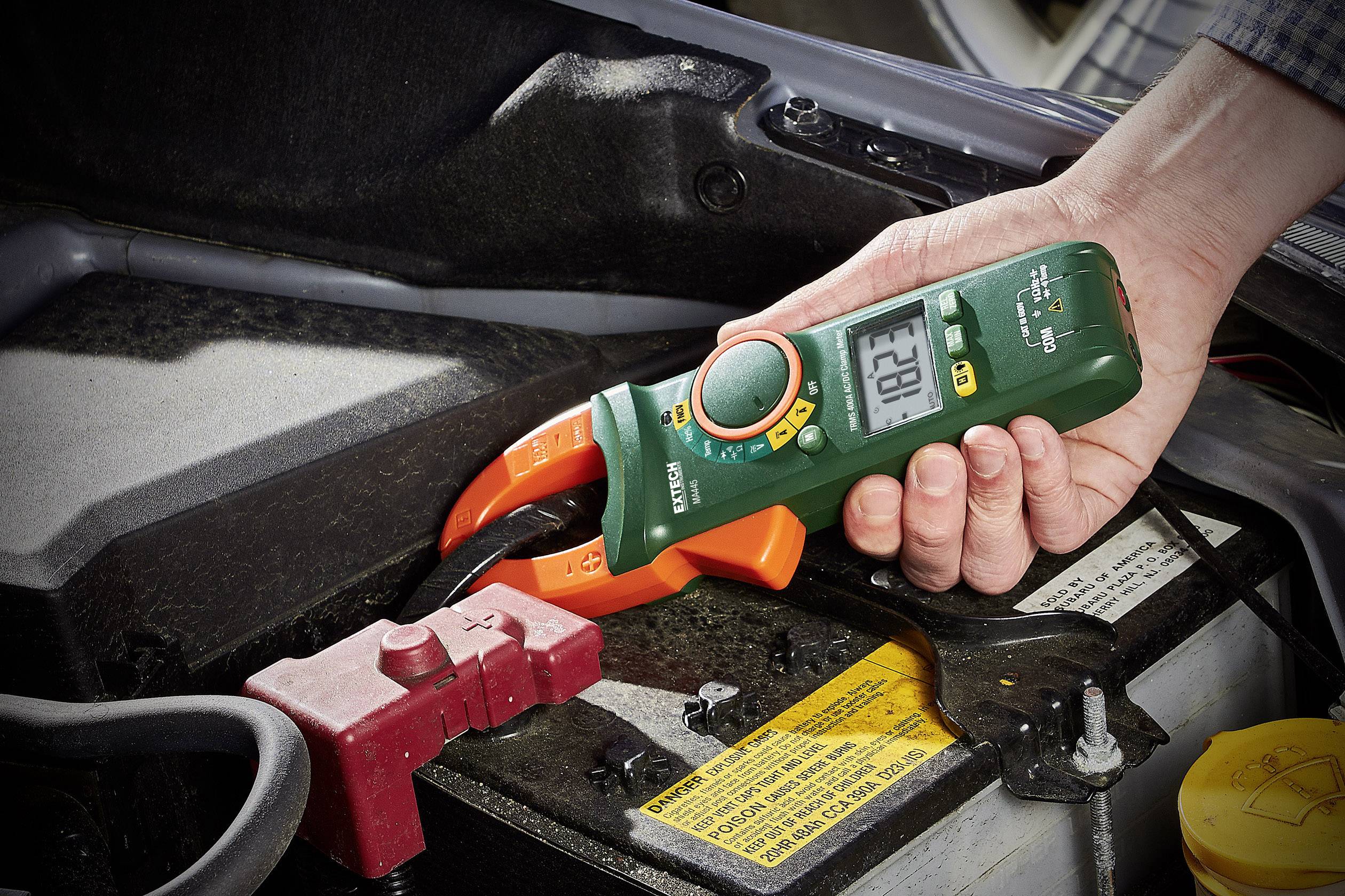 Ruka drží digitálny multimeter a meria napätie autobatérie pod kapotou. Displej zobrazuje hodnotu '12.6'.