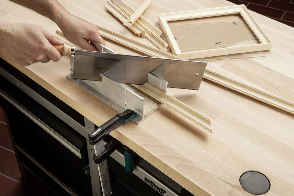 Na stolárskej lavici muž pílkou v pokosnici rozrezáva drevený rám. V pozadí sú viditeľné drevené lišty.