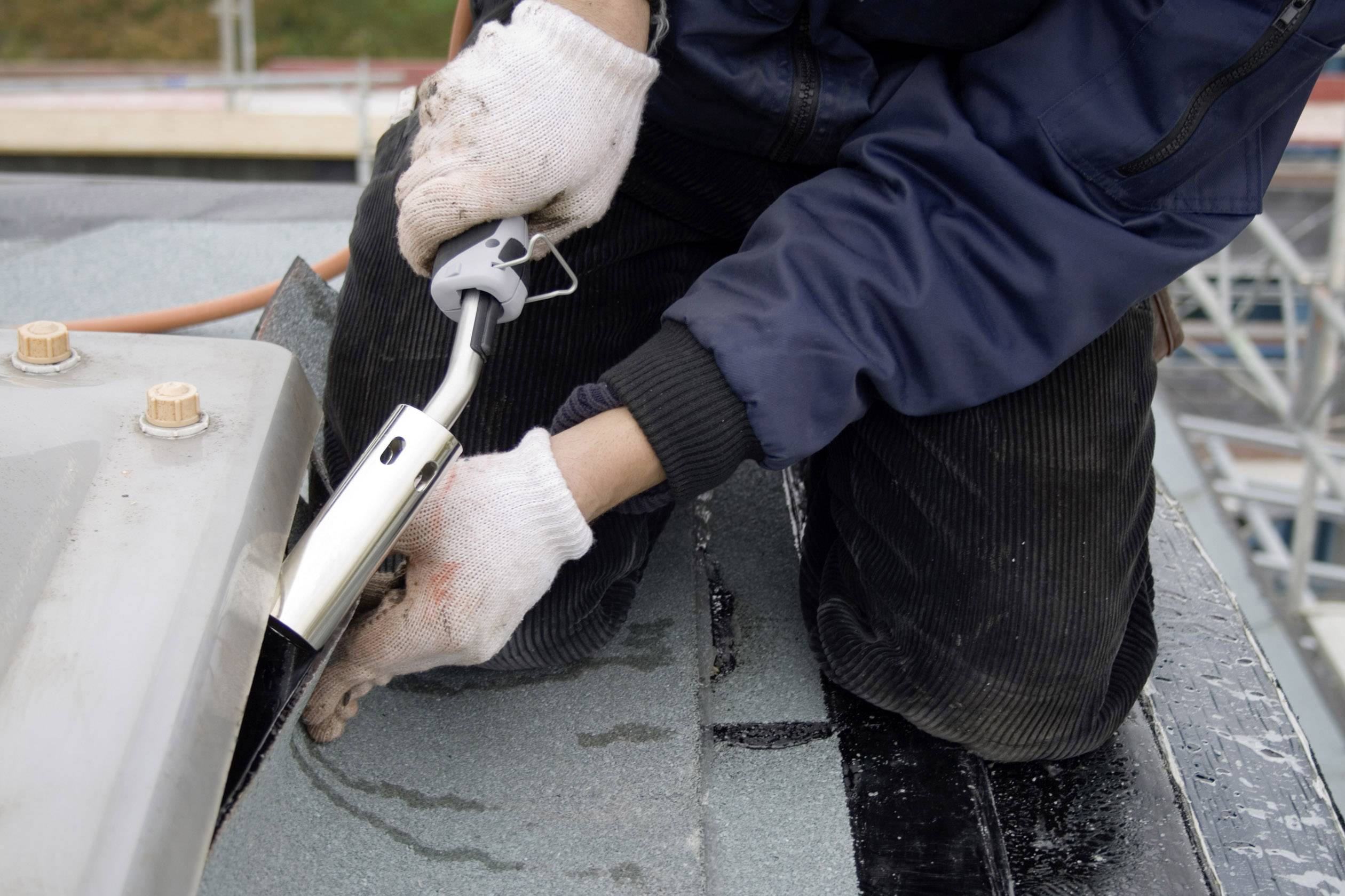 Osoba v bielych rukaviciach používa nástroj na spracovanie hydroizolačného materiálu strechy. Prostredie je stavenisko.