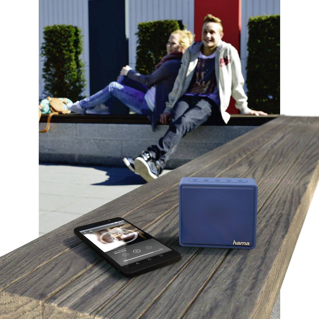 Pár sedí na lavičke; v popredí smartphone a modrý bluetooth reproduktor značky Hama.