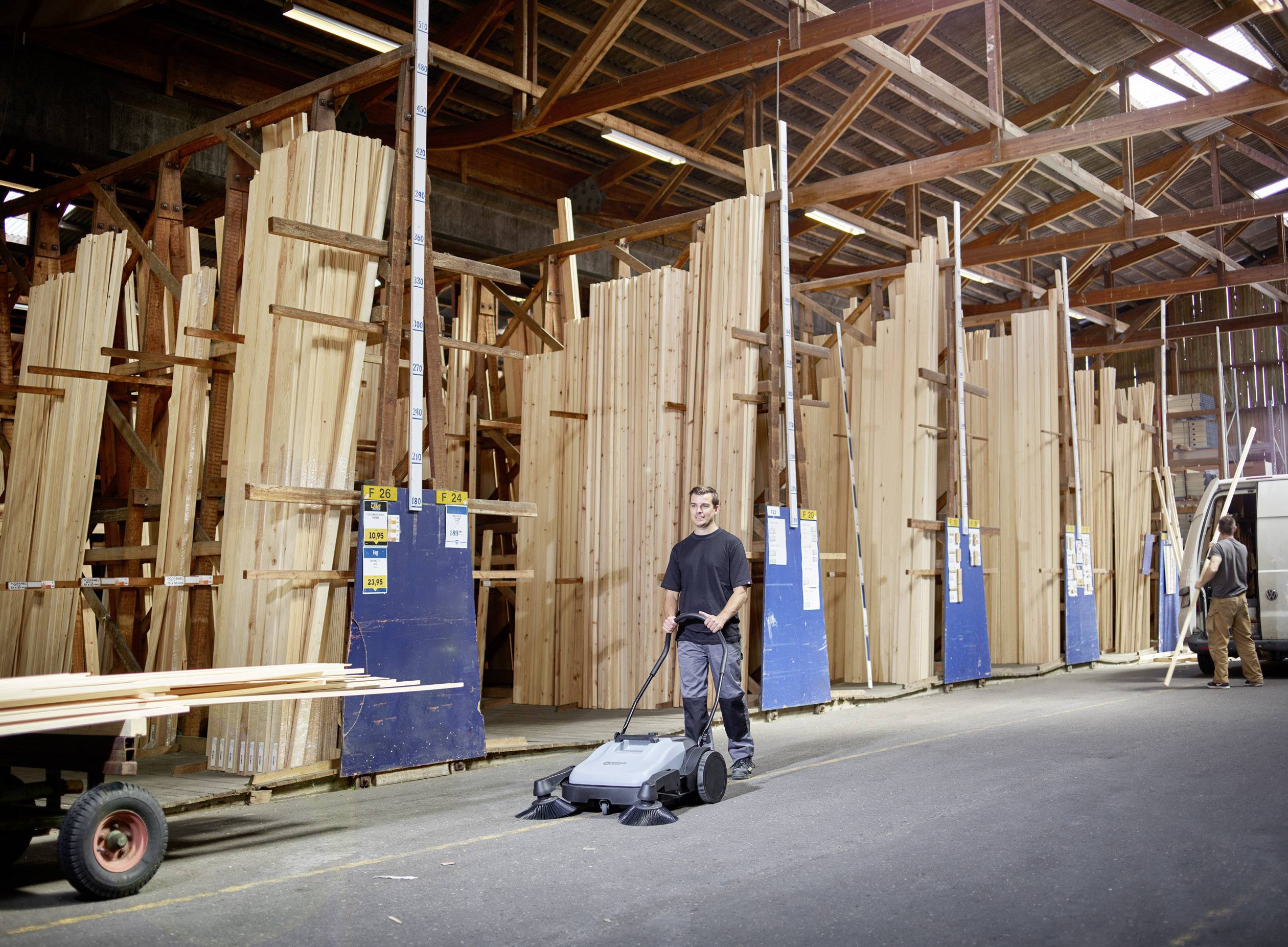 Osoba zametá podlahu veľkej drevárskej dielne. V pozadí sú vysoké stohy surových drevených dosiek.