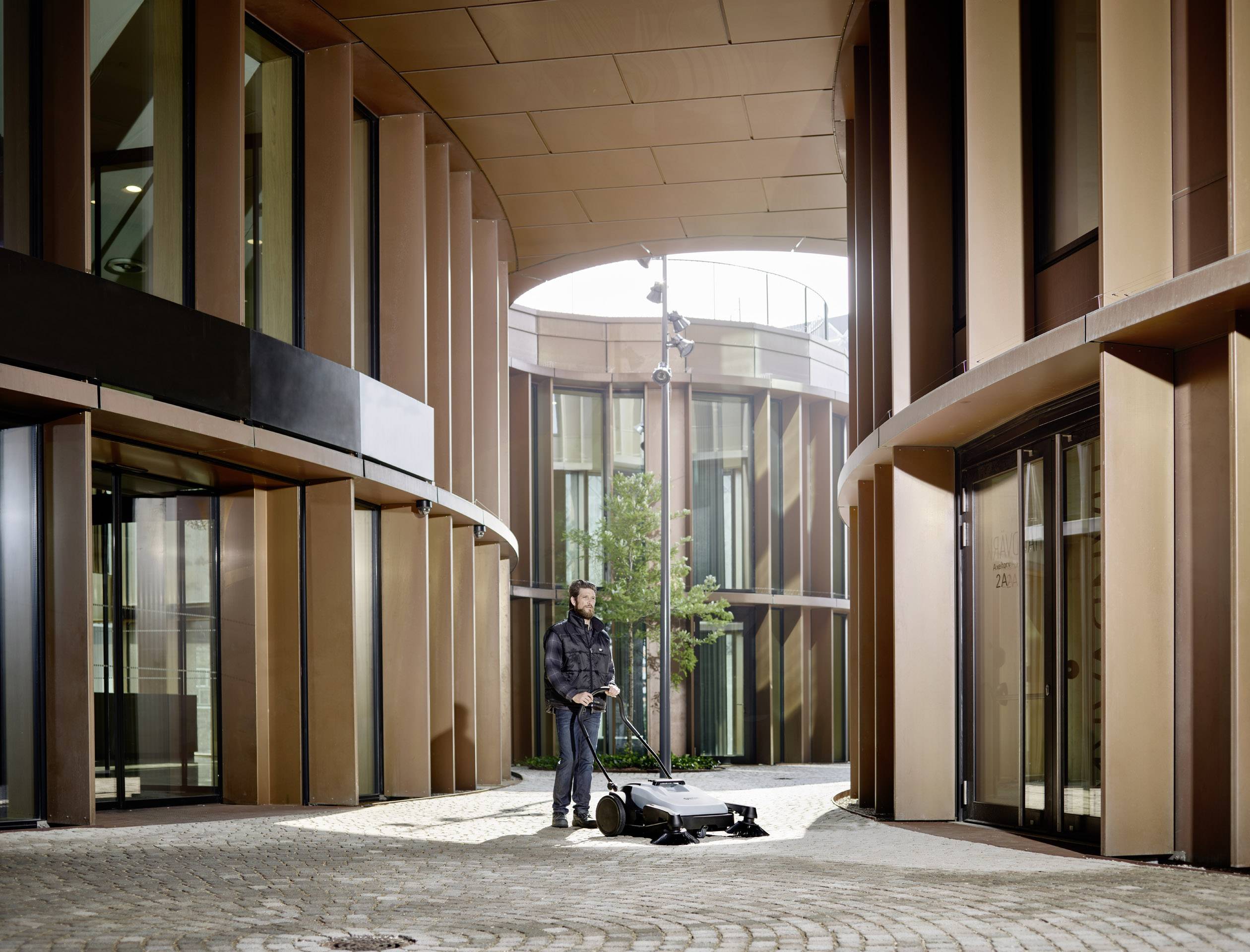 Osoba stojí s bicyklom v modernom, zakrivenom priechode medzi budovami s veľkými oknami a rastlinou v pozadí.