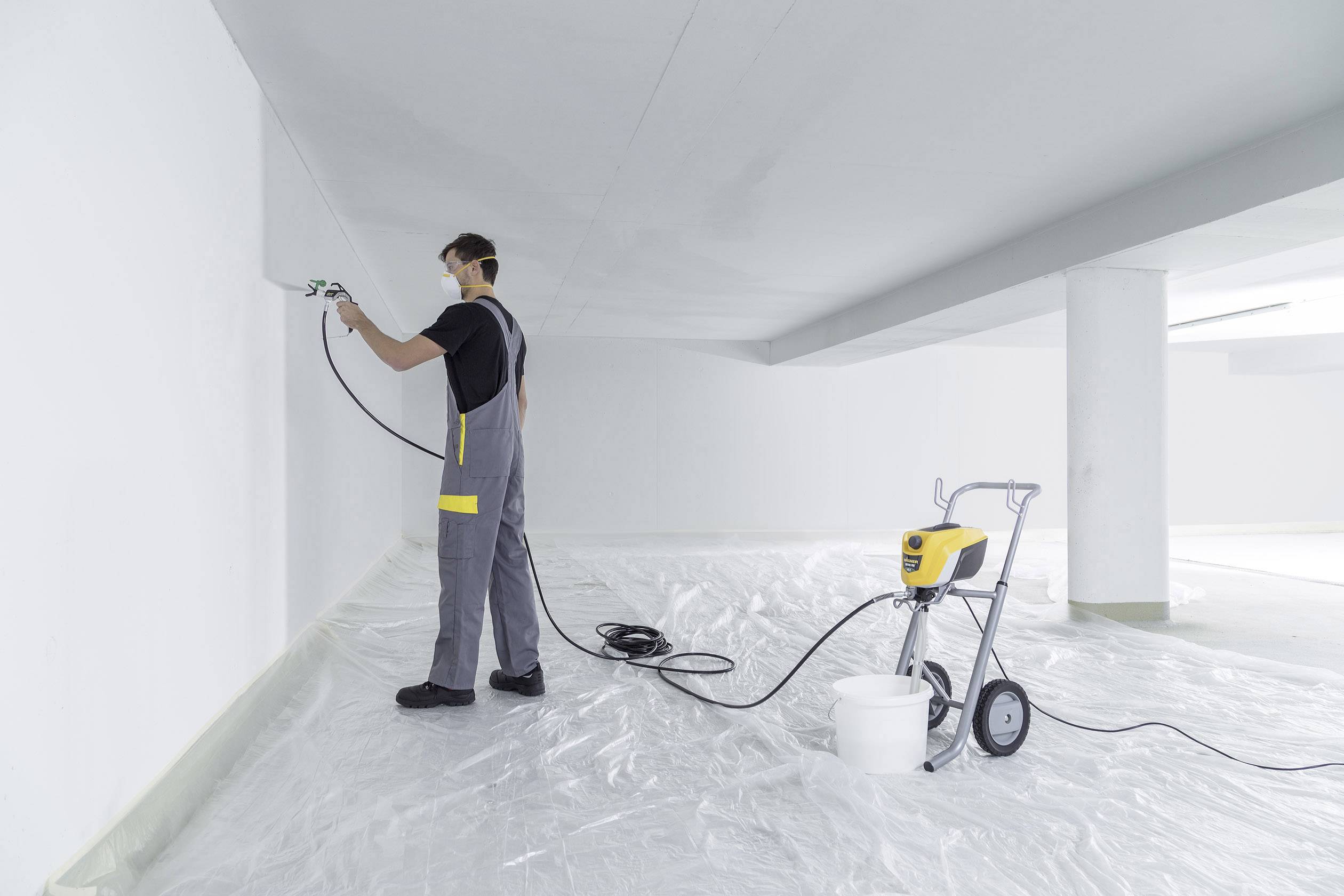 Robotník v ochrannom oblečení strieká pomocou stroja bielu farbu na stenu vo veľkej, prázdnej miestnosti.