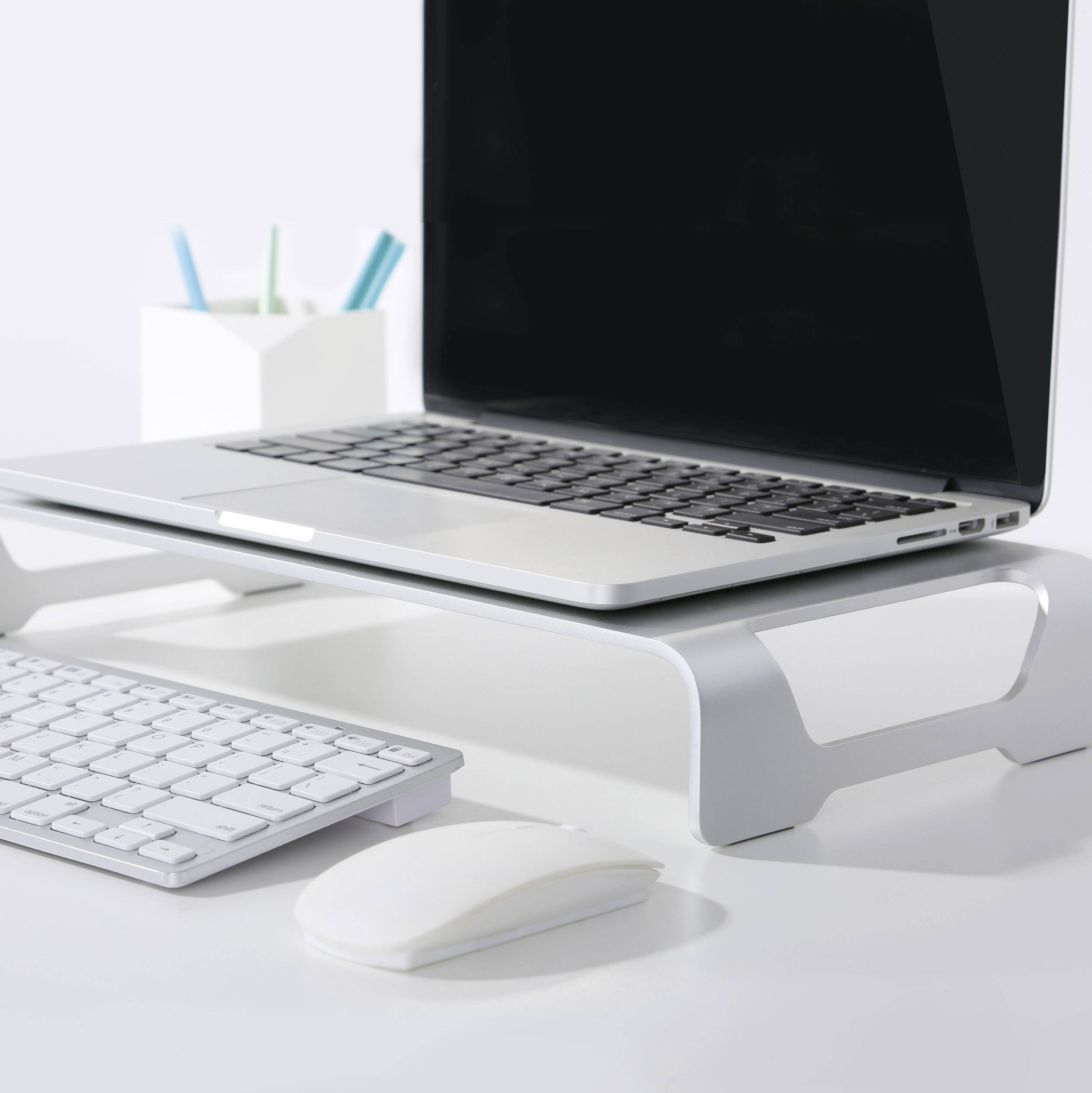 Laptop na zvýšenom stojane, vedľa bezdrôtová myš a klávesnica. V pozadí stojan na perá s perami.