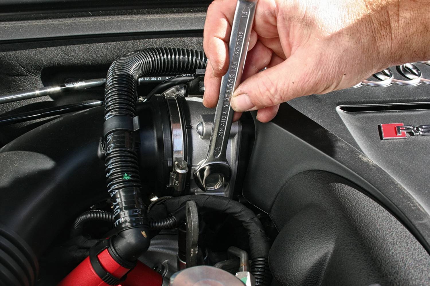 Jedna ruka drží kľúč a uťahuje skrutku v motorovom priestore automobilu. V pozadí sú viditeľné káble a potrubia.