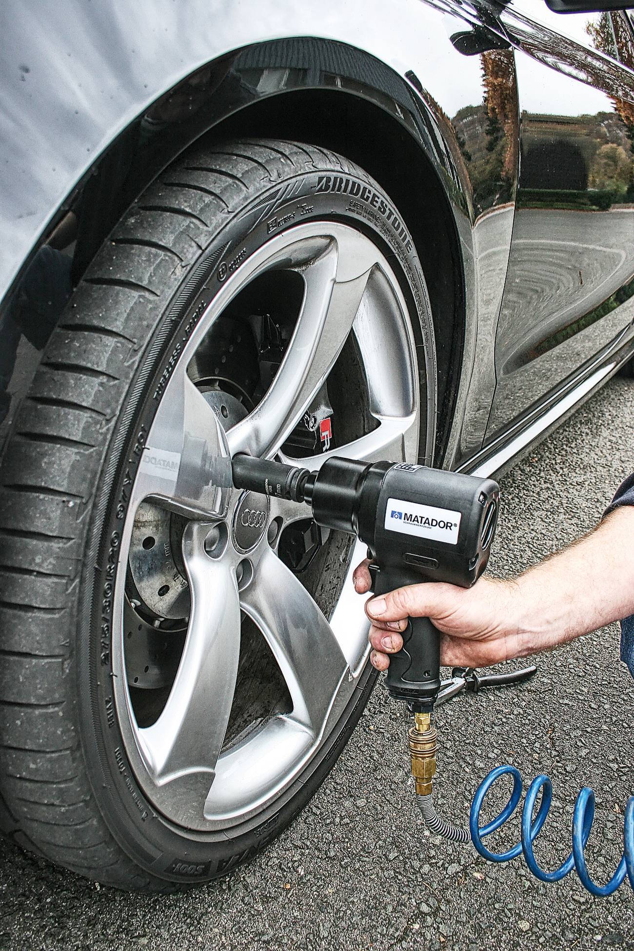 Jedna ruka drží pneumatický rázový uťahovák, ktorý pracuje na kolese automobilu.
