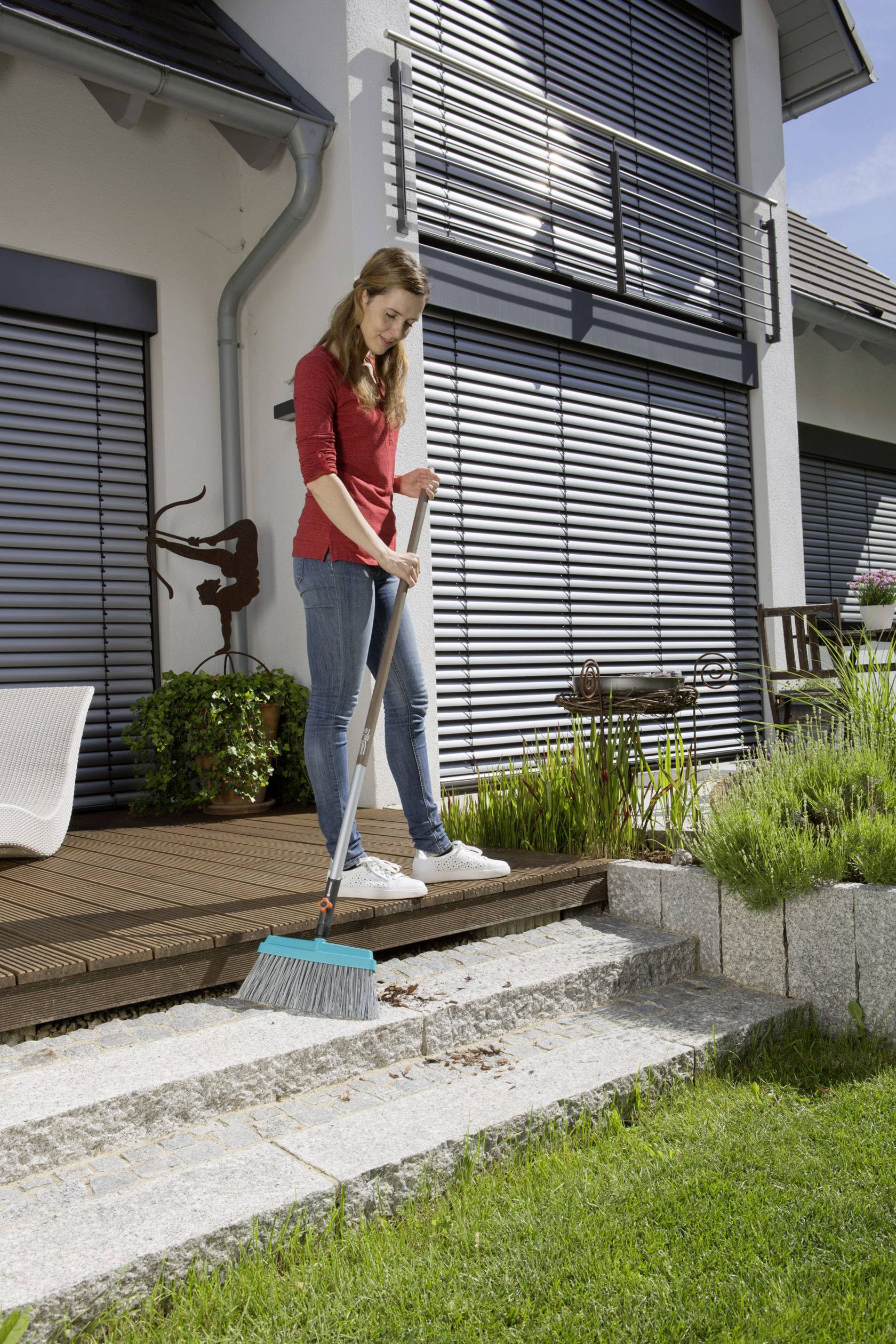 Žena zametá lístie z kamenného schodiska v modernej záhrade s terasou. V pozadí je dom so zatvorenými žalúziami.