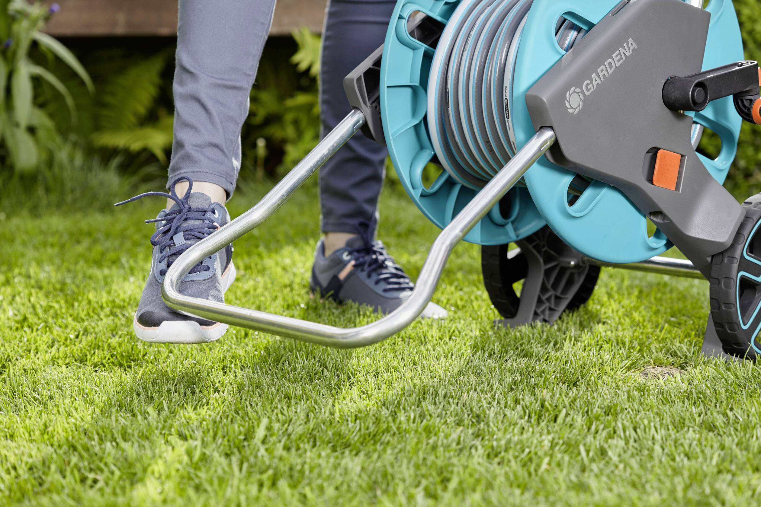 Osoba používa cievku na záhradnú hadicu na trávniku. Má na sebe športové oblečenie a pohybuje vozíkom s hadicou.
