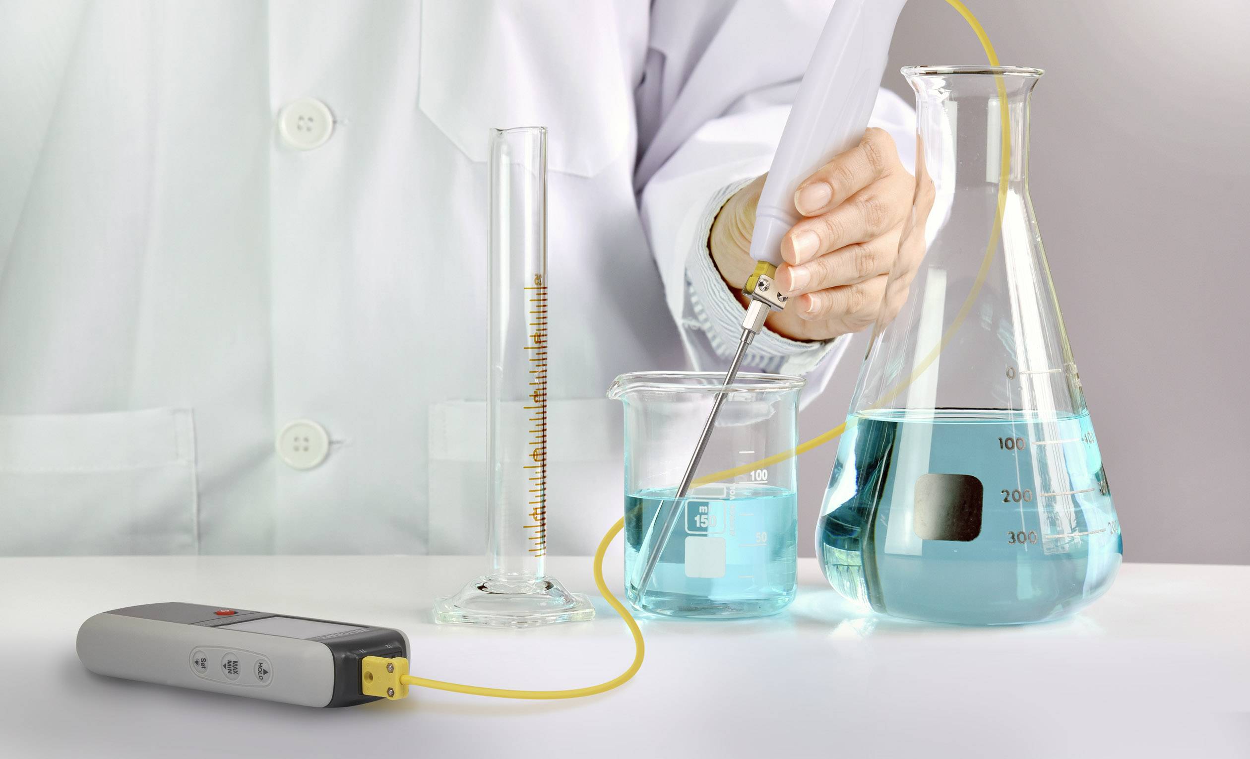 Osoba v bielom plášti meria pH hodnotu modrej kvapaliny v kadičke pomocou pH metra.