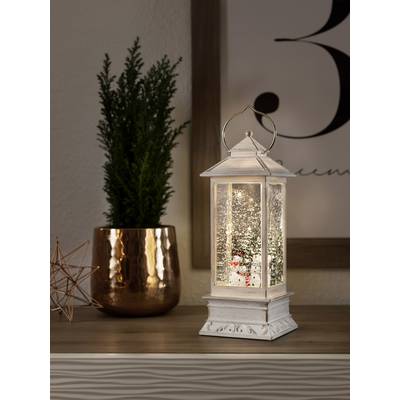 Biela lampa so snehuliakom ako dekoráciou, ktorá svieti na stole vedľa medeneného hrnca s malou rastlinou.