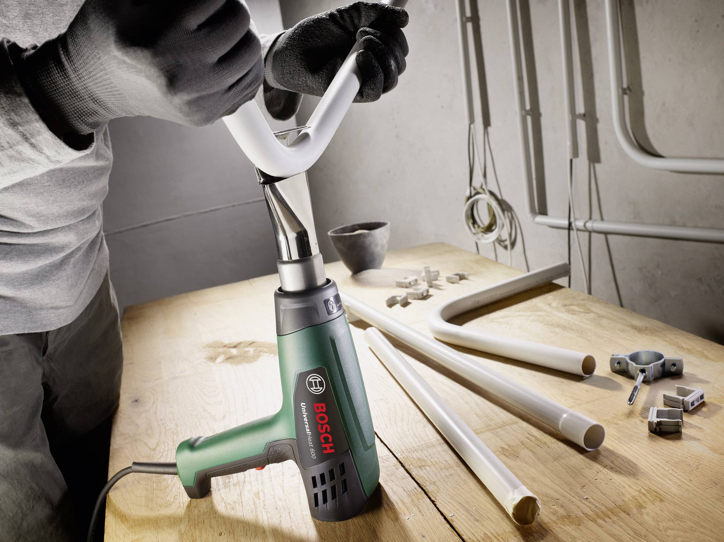 Osoba pracuje s teplovzdušnou pištoľou Bosch na bielej trubke na drevenom stole. V pozadí sú viditeľné ďalšie nástroje a trubky.
