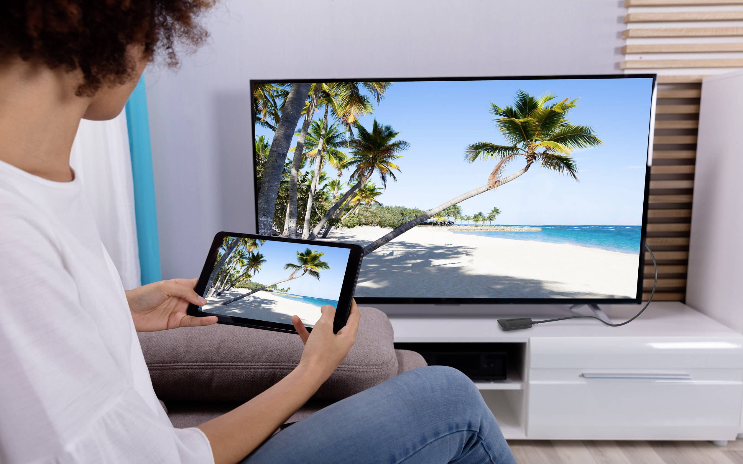 Osoba pozerá pláž s palmami na televízore, pričom drží to isté zobrazenie na tablete.