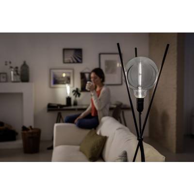 Žena sedí na pohovke v útulne osvetlenej miestnosti a pije z hrnčeka. V popredí stojí moderná stojanová lampa.