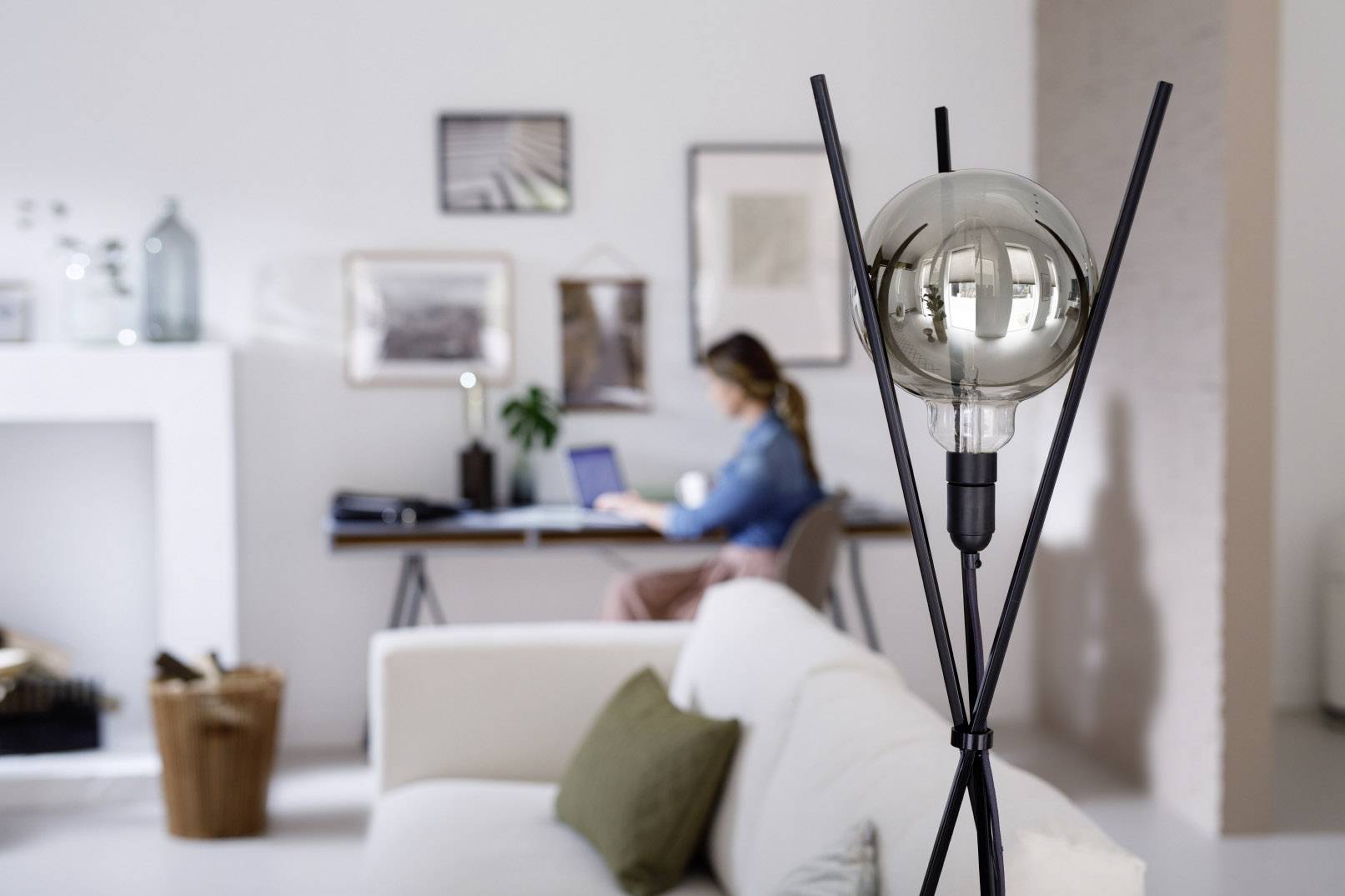 Lampa v popredí, rozmazaná osoba v pozadí pri stole s laptopom, svetlá miestnosť s obrazmi na stene.