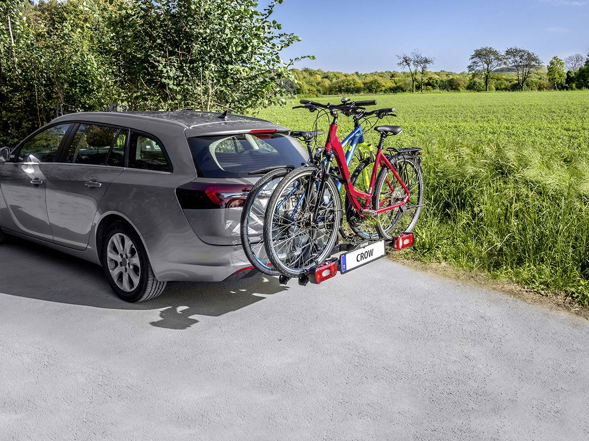Auto so strešným nosičom bicyklov a tromi bicyklami, zaparkované na vidieckej ceste. V pozadí zelené polia a stromy.