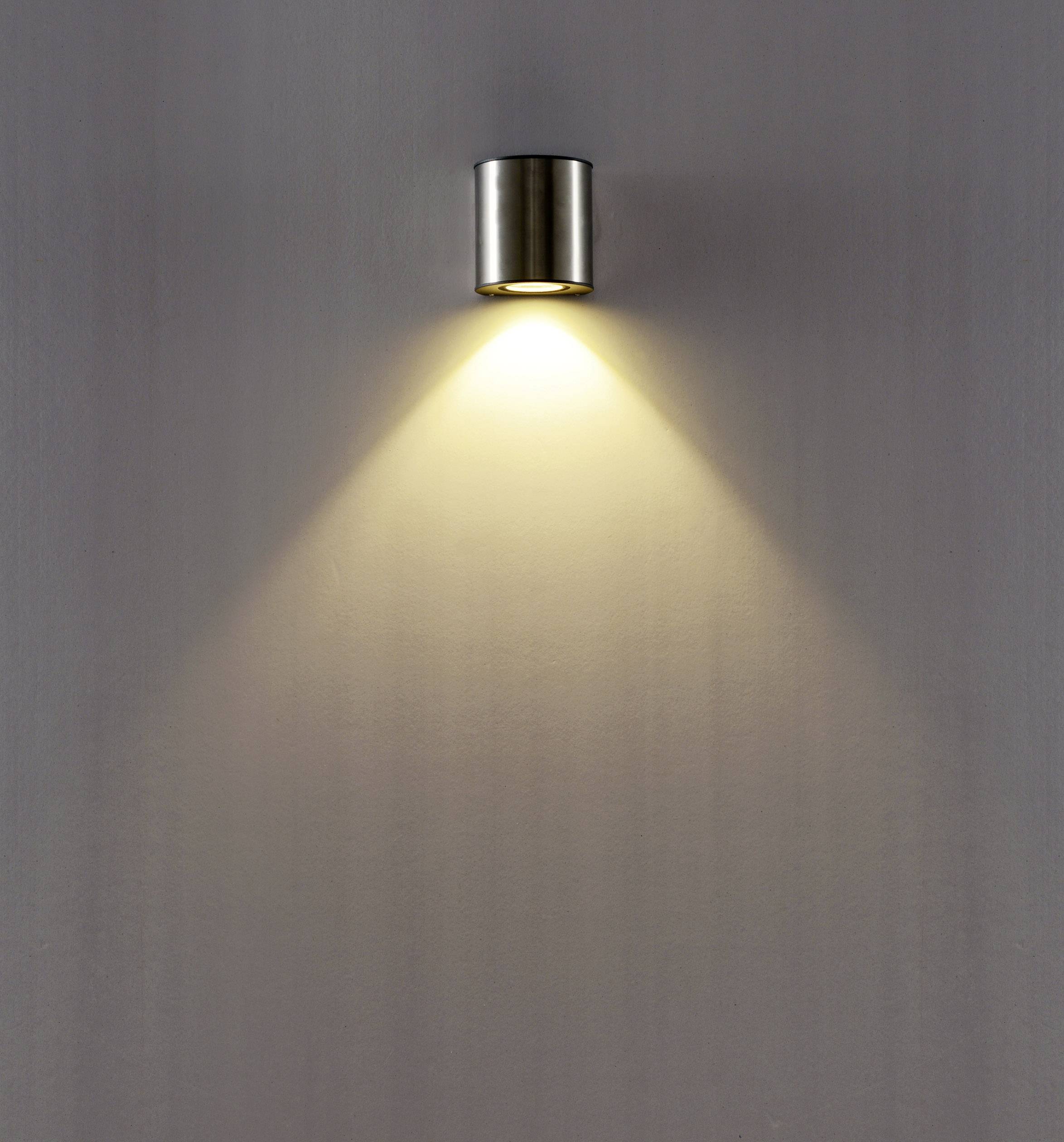 Kovená nástenná lampa s teplým svetelným lúčom, ktorý sa rozteká smerom nadol na hladkú stenu. Minimalistický dizajn.