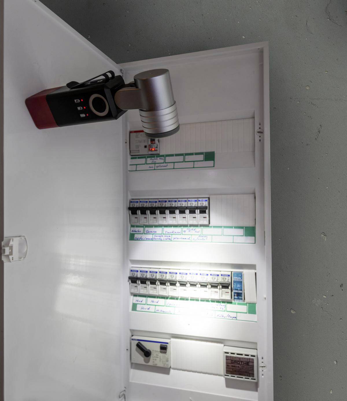 Otvorená elektrická rozvodná skriňa s viacerými poistkami a vypínačom. Baterka je nasmerovaná na rozvodňu.
