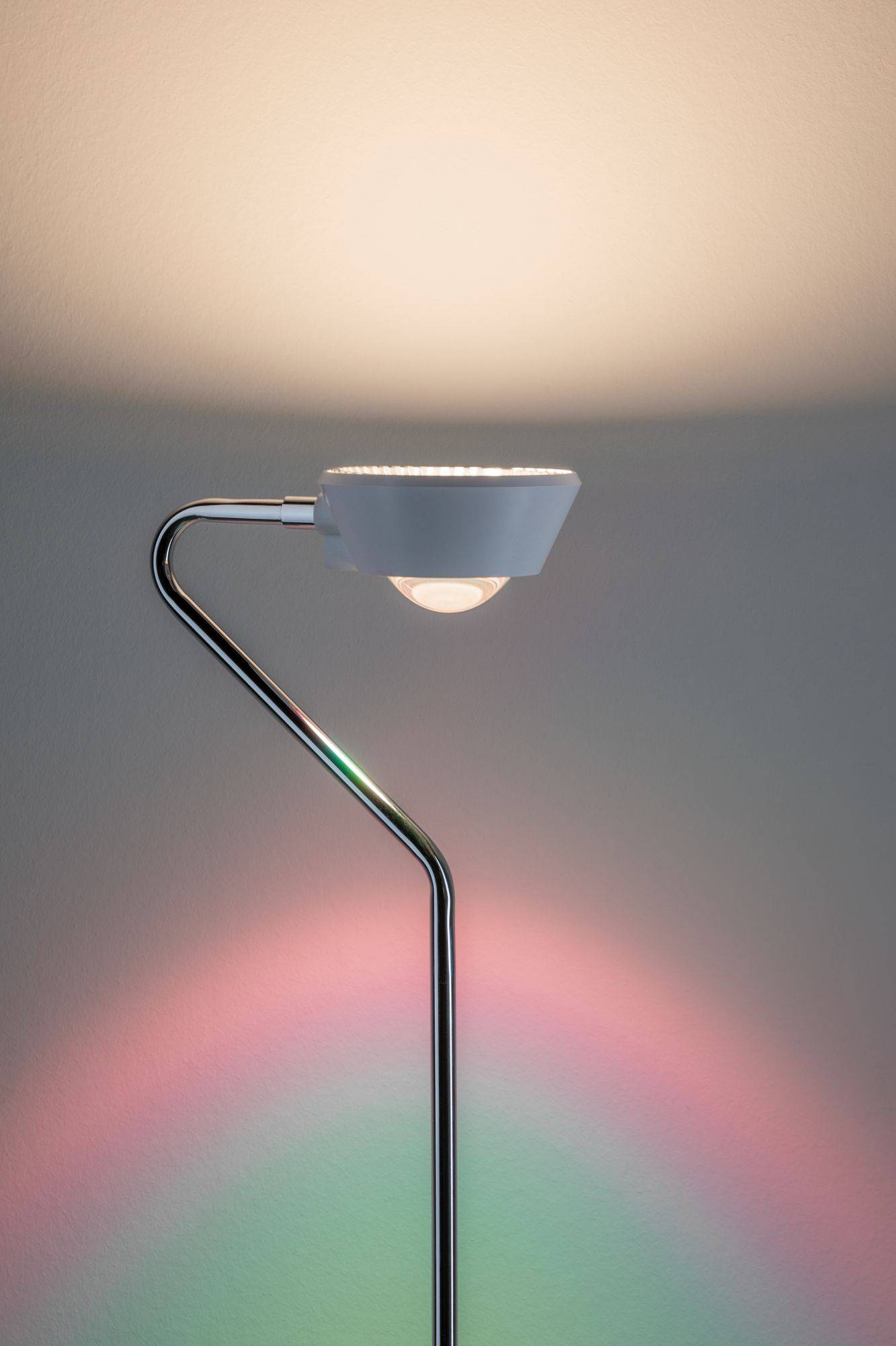Moderná stojaca lampa s oblúkovitou kovovým podstavcom vyžaruje jemné svetlo, ktoré na stenu premietá jemný dúhový efekt.