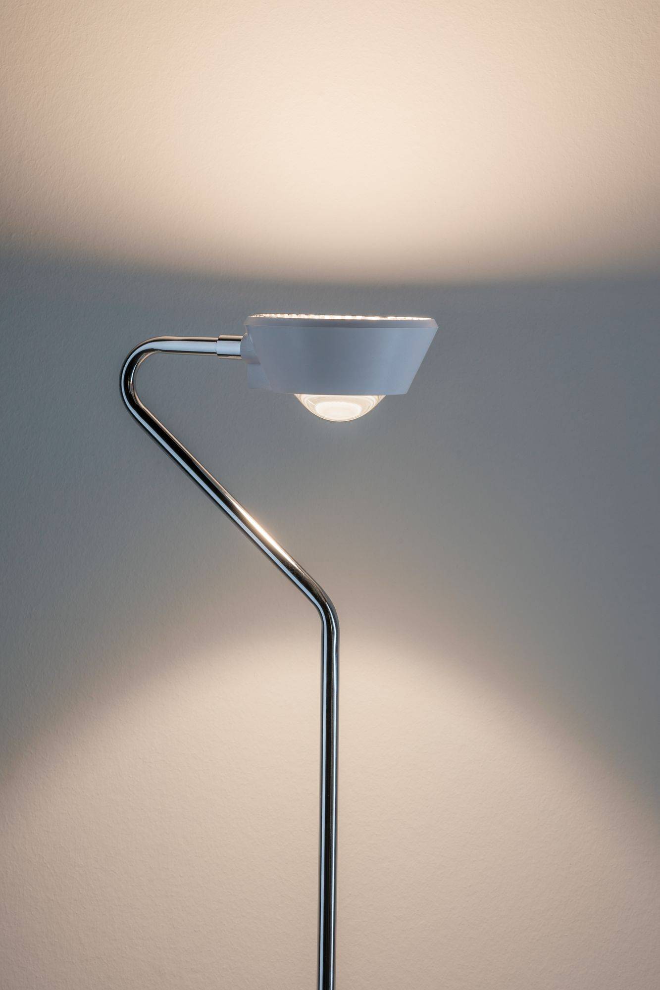 Moderná stojaca lampa so zakrivením kovovým rámom hádže mäkké svetlo na bielu stenu.