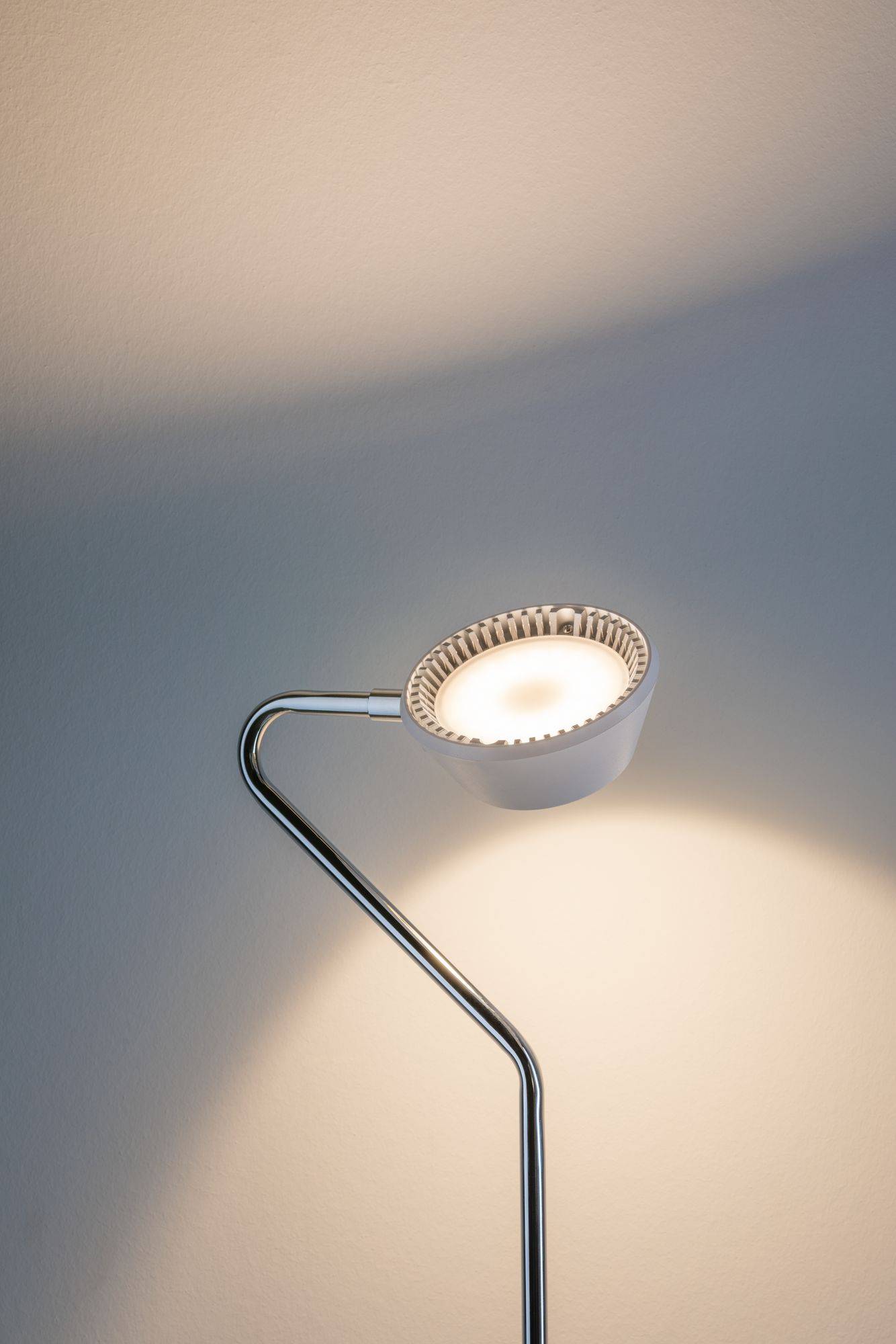 Moderná, minimalistická stojanová lampa svieti oproti stene. Lampa má kovový, zakrivený ramený a guľatý tienidlo.