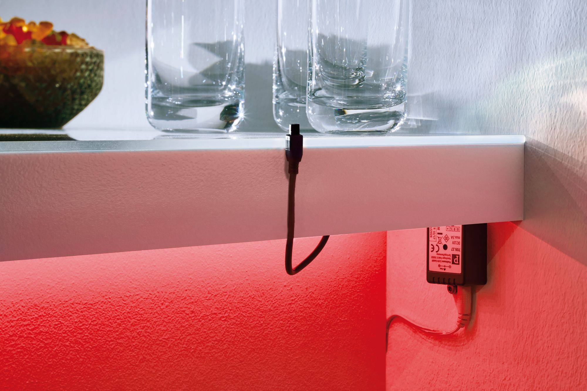 Polica so sklenenými nádobami a misou, pod ktorou trčí kábel od LED transformátora. Pozadie je osvetlené červeným svetlom.