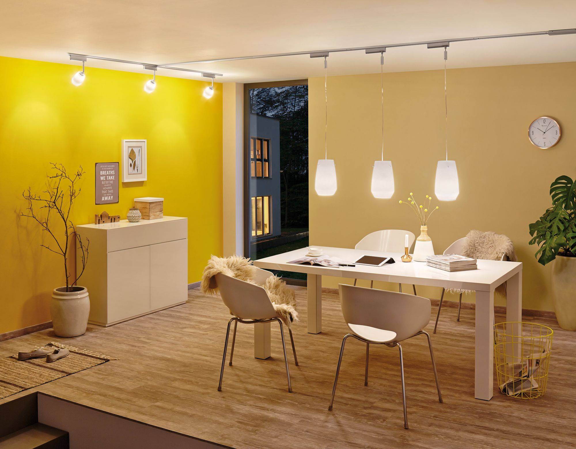 Moderne zariadená jedáleň so žltými stenami, bielym stolom, stoličkami a štýlovými lampami. Veľké okná poskytujú výhľad von.
