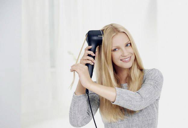 Žena s dlhými blonďavými vlasmi používa čierny styling nástroj na vlasy. Má na sebe sivý sveter a usmievavo pôsobí uvoľnene.