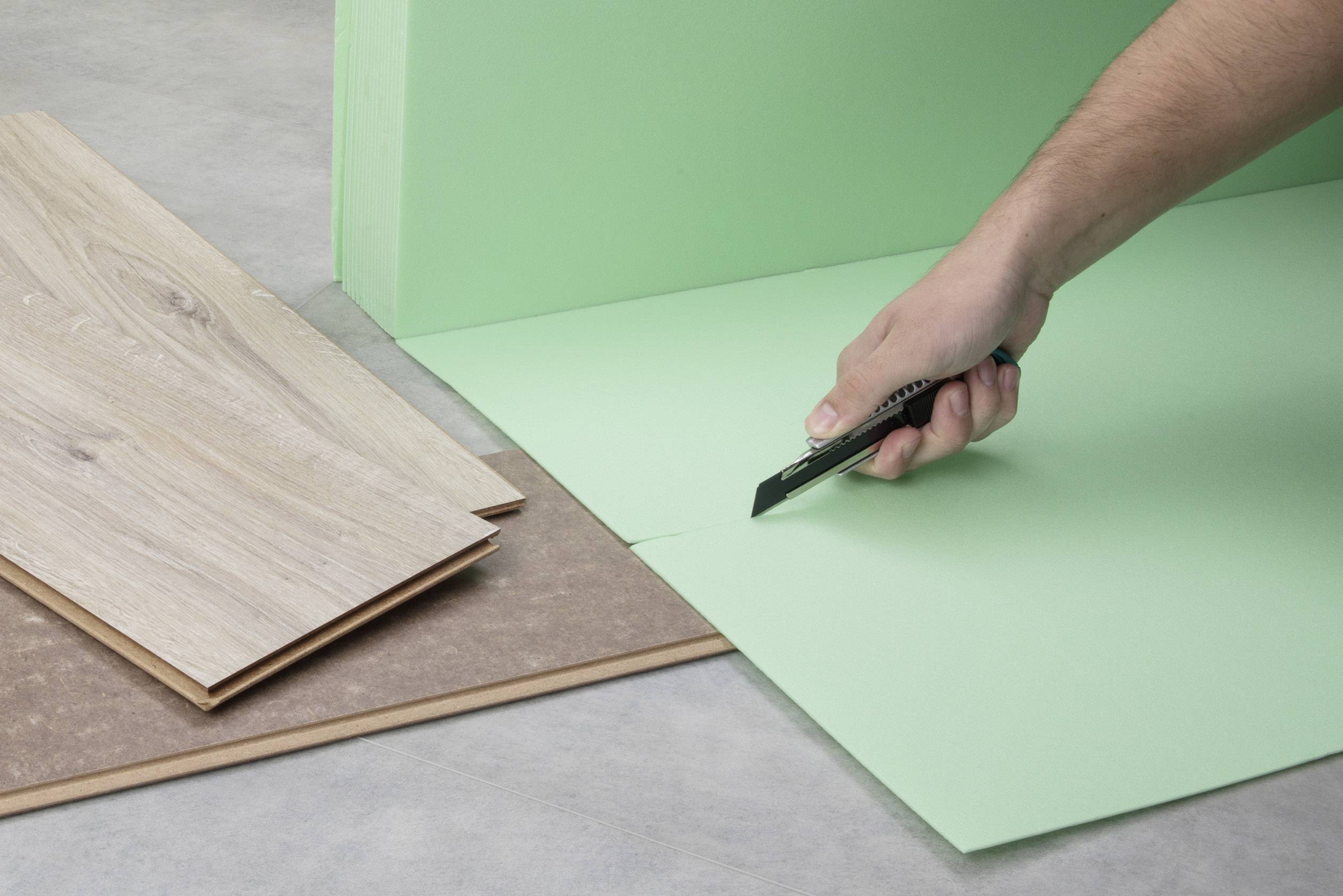 Ruka krája rezacím nožom zelenú podložku pre laminátové podlahy. Vedľa sú pripravené laminátové dosky.