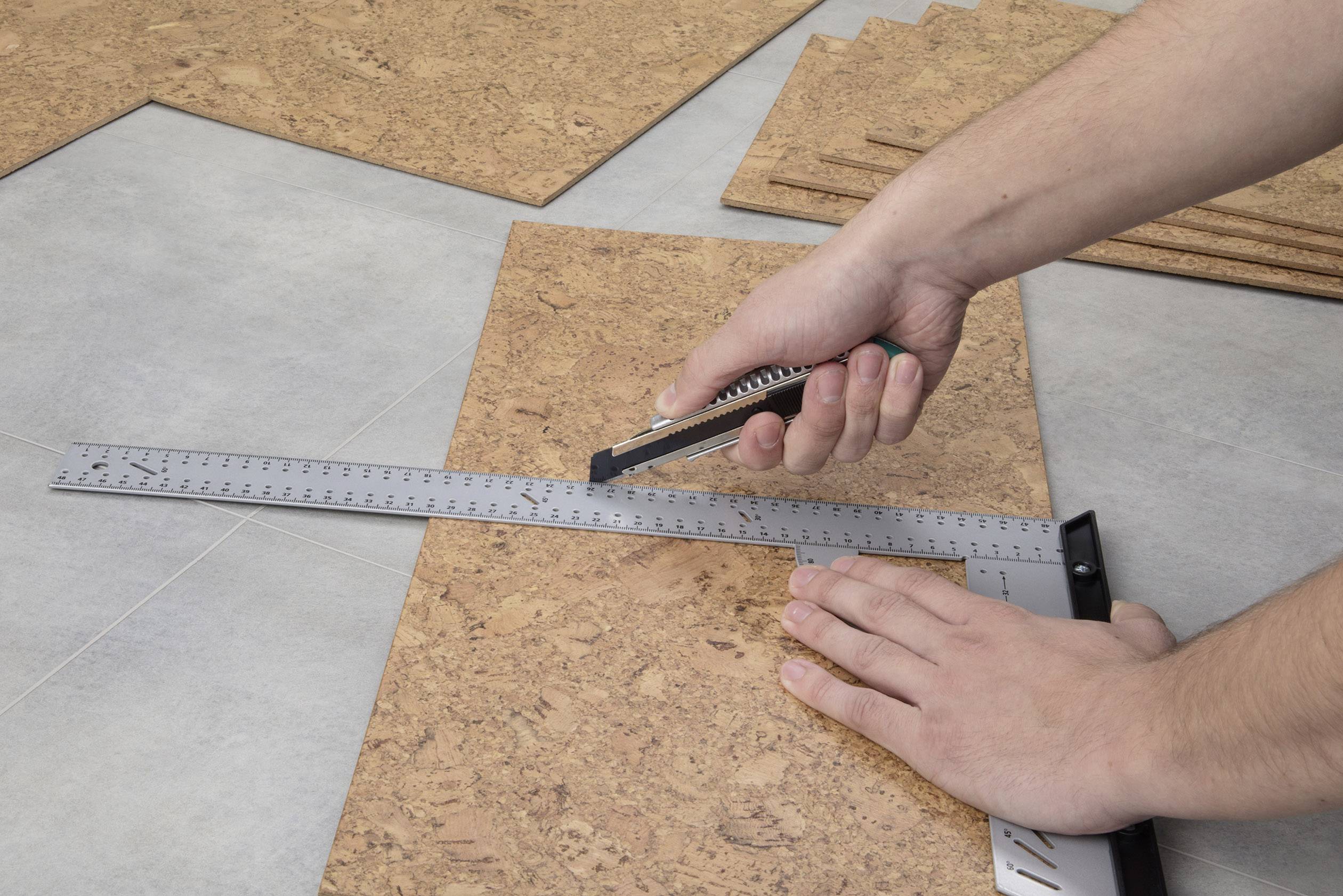 Osoba krája korkový dosku pomocou odlamovacieho noža, podporovaná kovovým pravítkom na sivej podlahe.