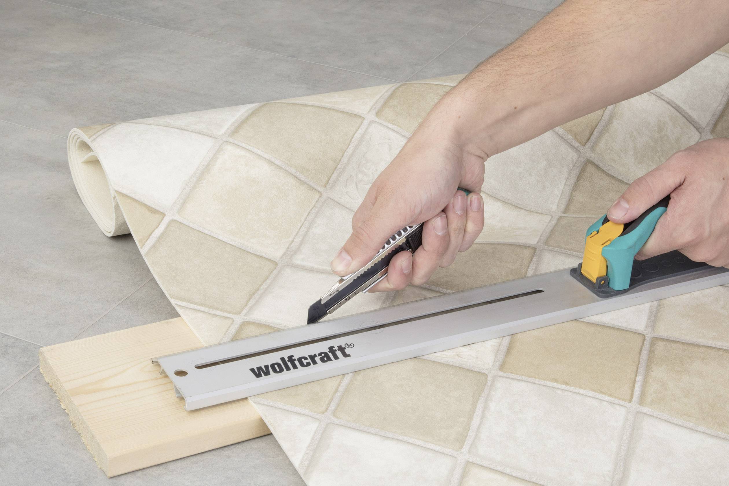 Osoba strihá béžovú dlažbu pomocou odlamovacieho noža a kovovej pravítka na drevenej doske.