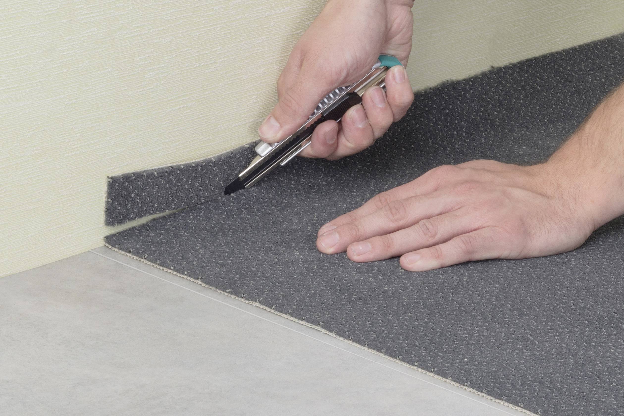 Osoba krája sivý koberec kobercovým nožom, ktorý je položený na podlahe. Ruky sú blízko pri koberci.