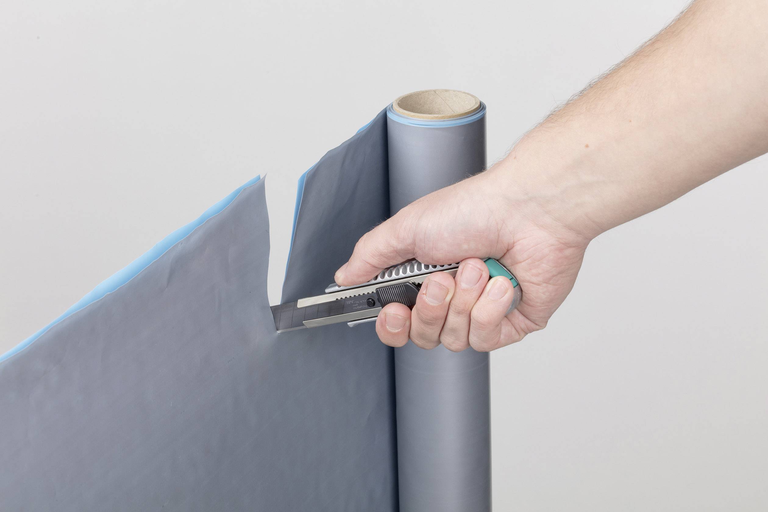 Jedna ruka krája priemyselnou rezačkou sivú plastovú fóliu z role. Fólia je čiastočne odrezaná.