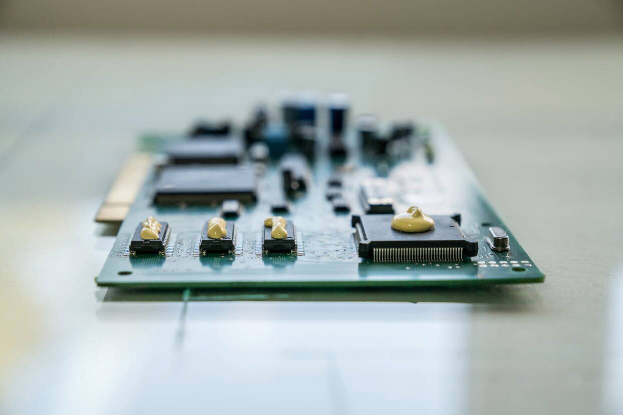 Detailný záber zelenej dosky plošných spojov s viacerými elektronickými komponentmi namontovanými na jej povrchu, ležiacej na stole.