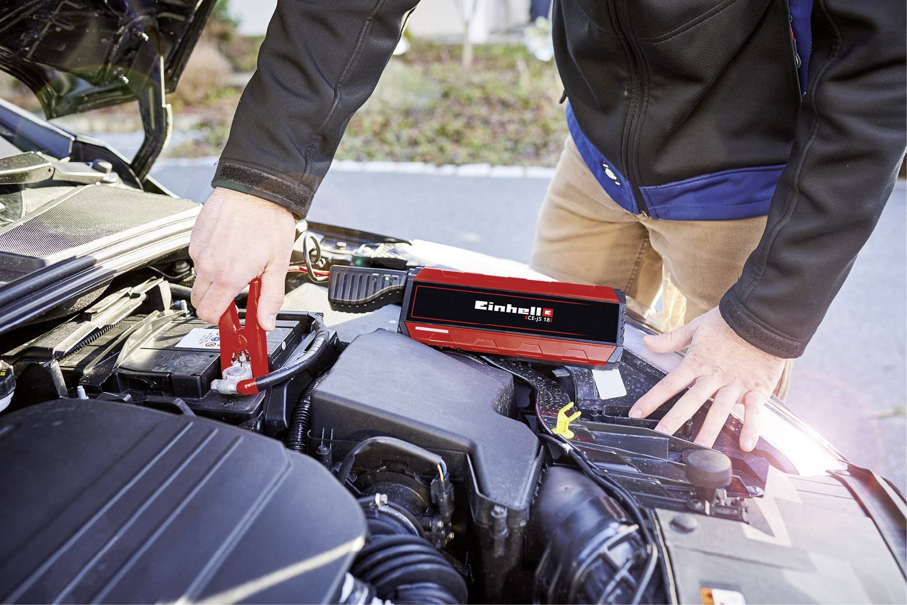Muž používa prenosný štartovací pomocník na naštartovanie auta. Zariadenie je červené a leží na bloku motora.