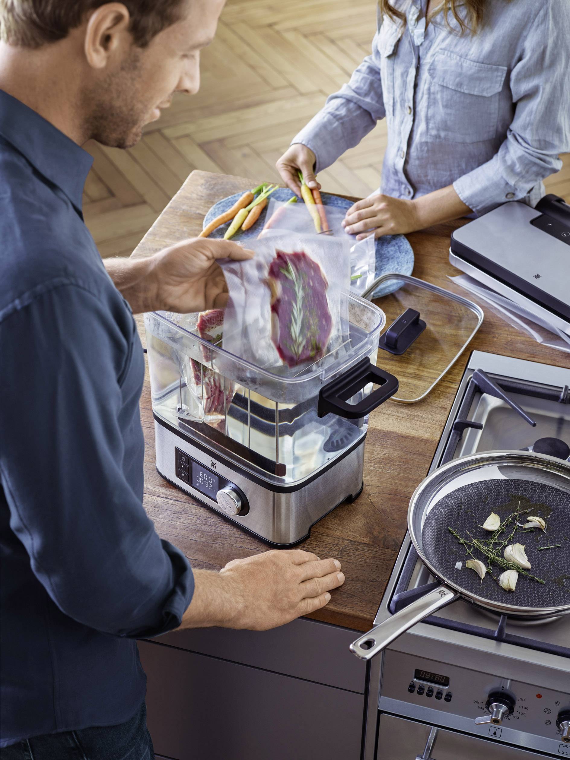 Muž vkladá vákuovo balené mäso do parného hrnca. Žena lúpe mrkvu. Na sporáku stojí panvica s bylinkami.