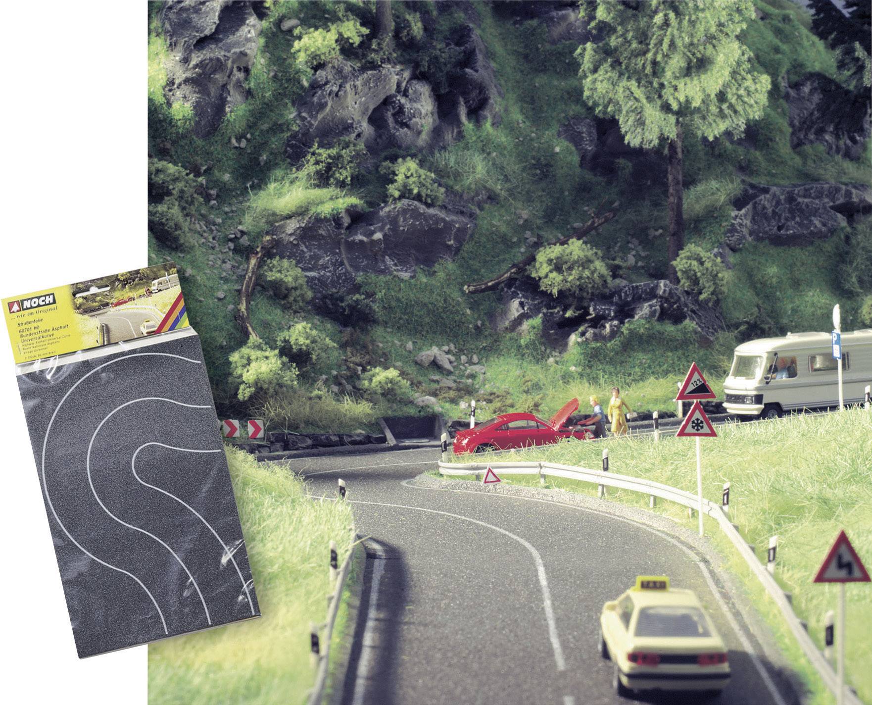 Model'ová cesta s krivkami, červené auto vedľa cesty, obytný príves v pozadí. V popredí je viditeľný živý plot.