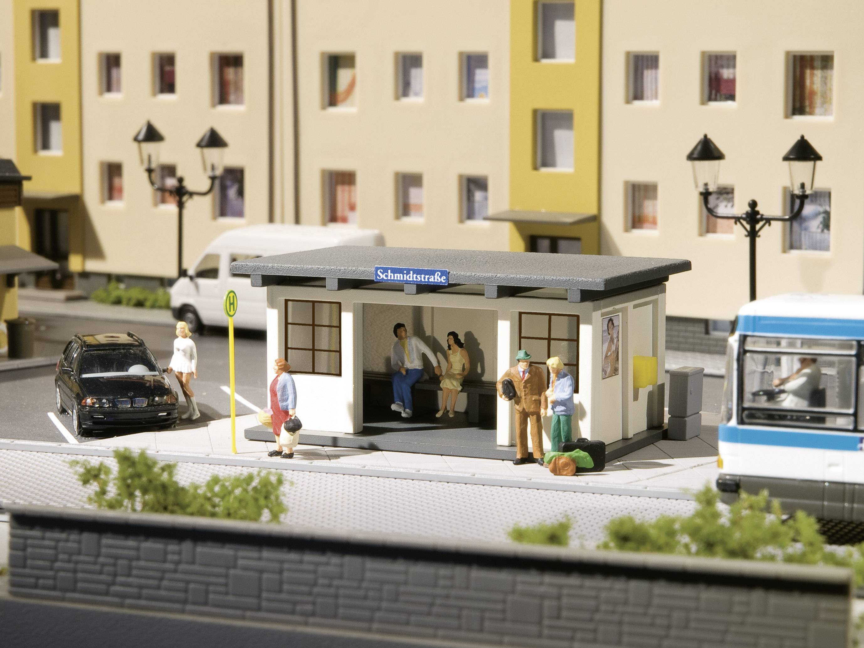 Zastávka v miniaturnej mierke na ulici 'Kováčska' s čakajúcimi figúrkami. Budovy v pozadí, auto a autobus sú pripravené.